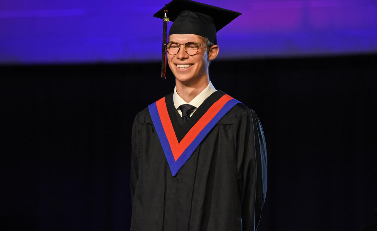 A student wearing cap and gown smiles on stage during graduation.