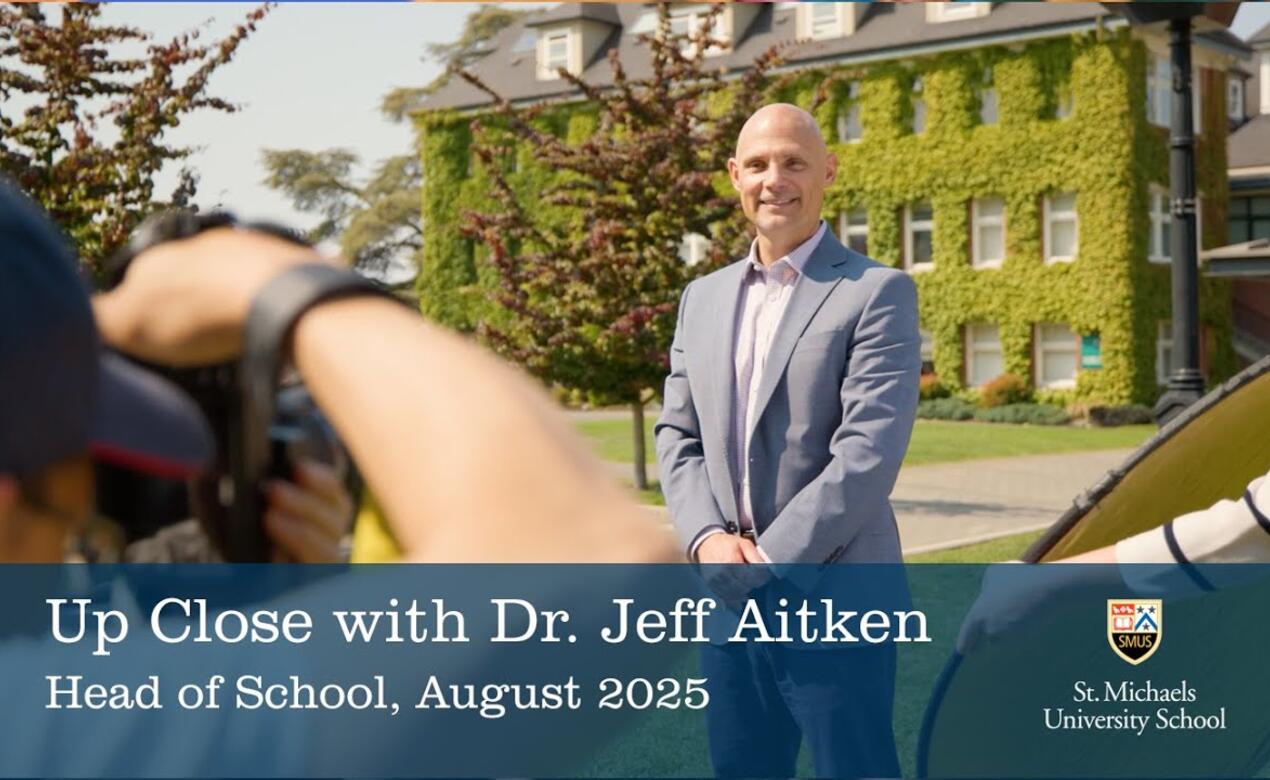 Dr. Jeff Aitken poses for a photo with the School House Building in the background and the photographer in the foreground.