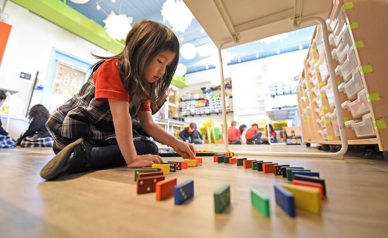 A Kindergarten student lines up dominoes while sitting underneath a table in the Imagination Lab.