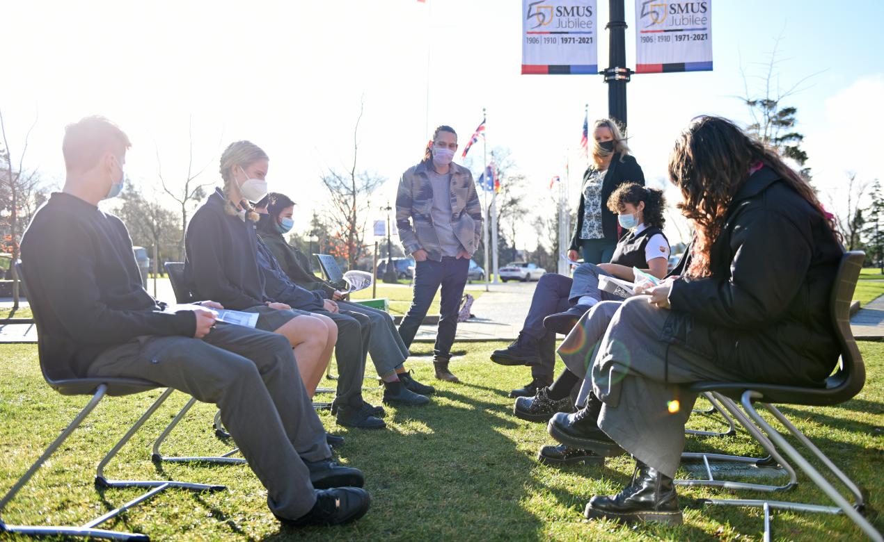 Salish artist Dylan Thomas works with Senior School math students outside while explaining the bone game.