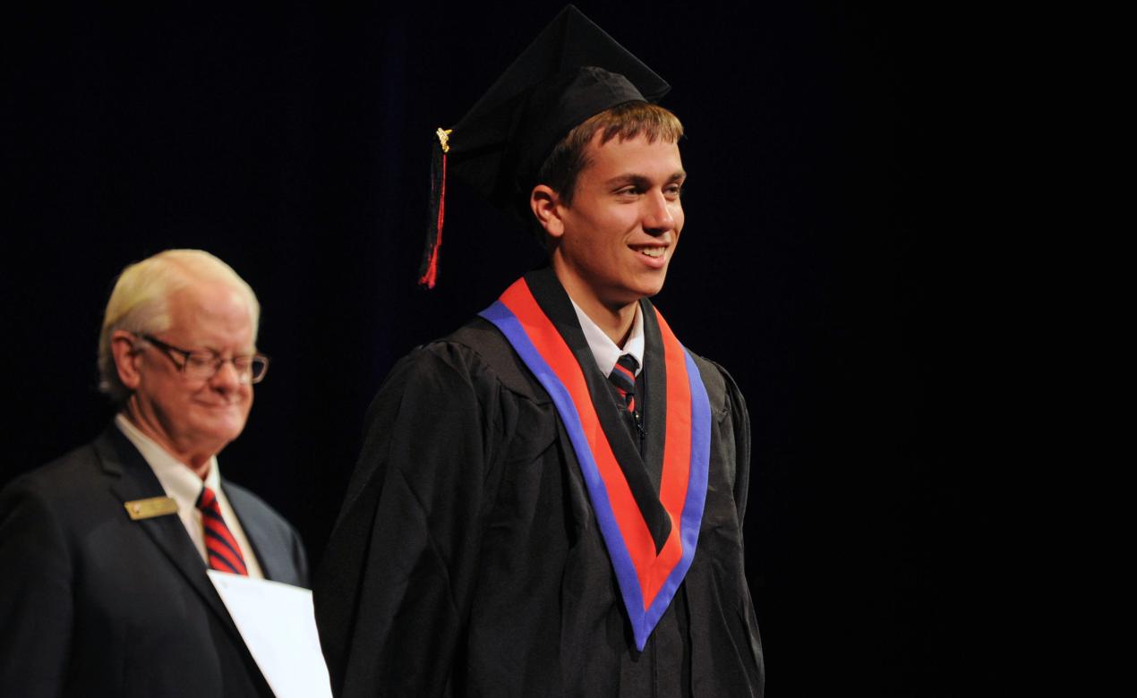 A student wearing a cap and gown smiles on stage during graduation