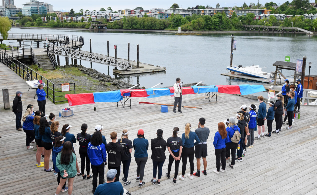 An aerial photo of group of rowers standing on a dock in a semicircle around a covered boat and a speaker.