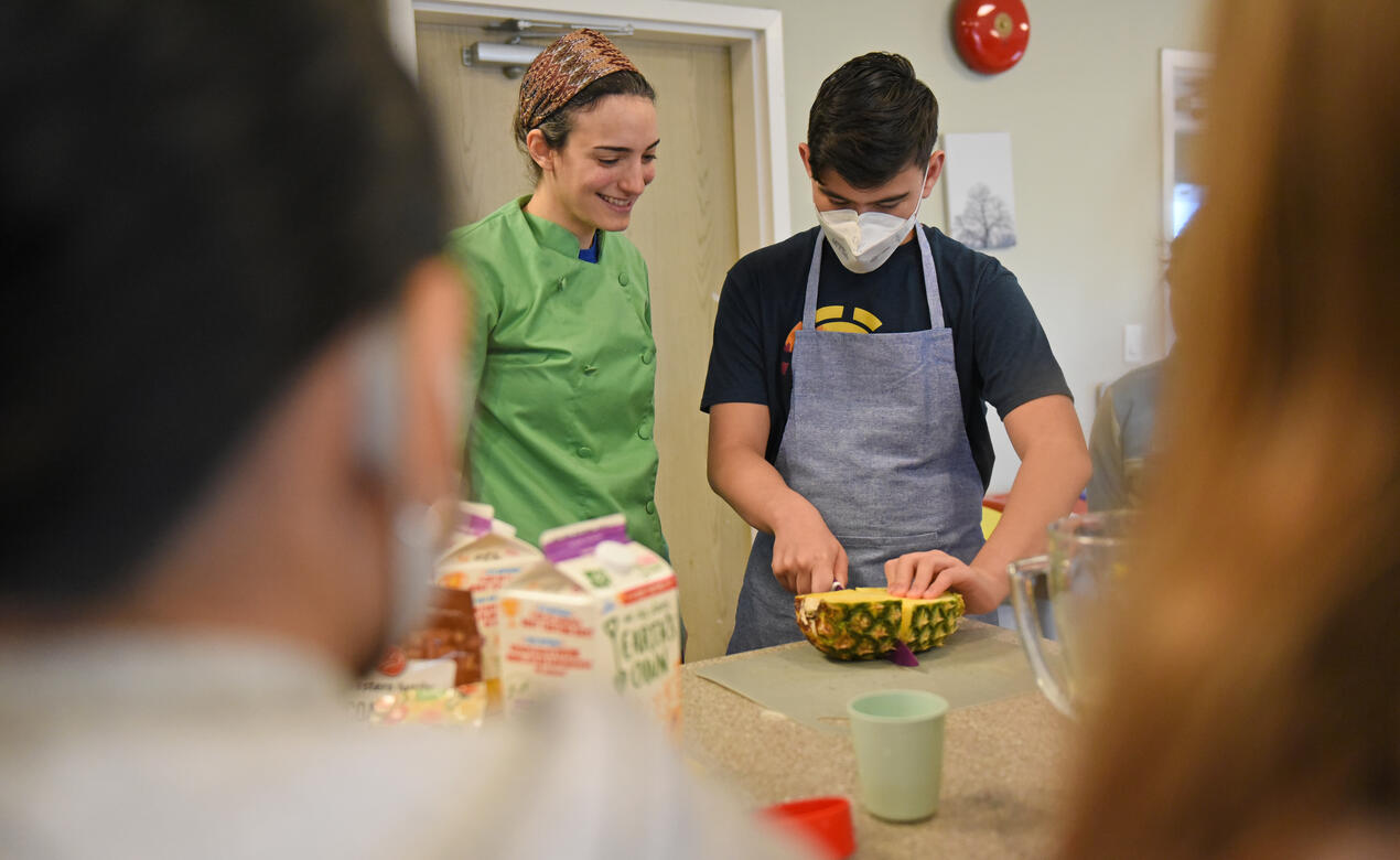 An instructor watches as a student cuts pineapple during a cooking class in the boarding house kitchen.