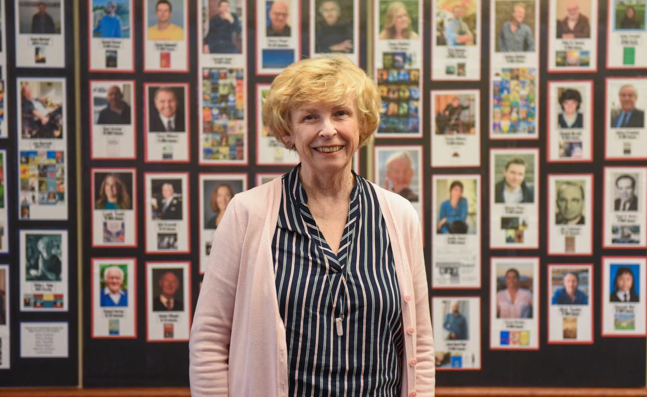Joan Tweedie, Senior School librarian, stands in front of a display wall that features photos of alumni and book covers.