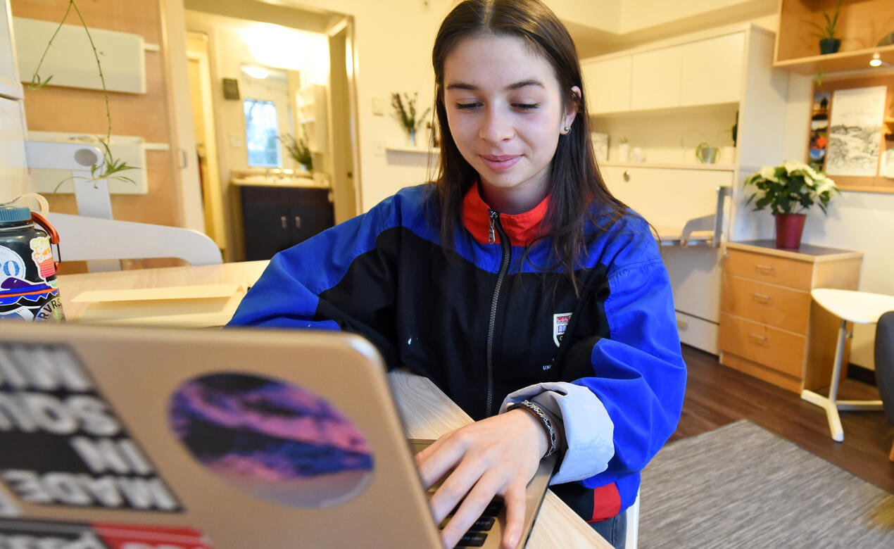 A student sits at a desk on their laptop in their boarding room.