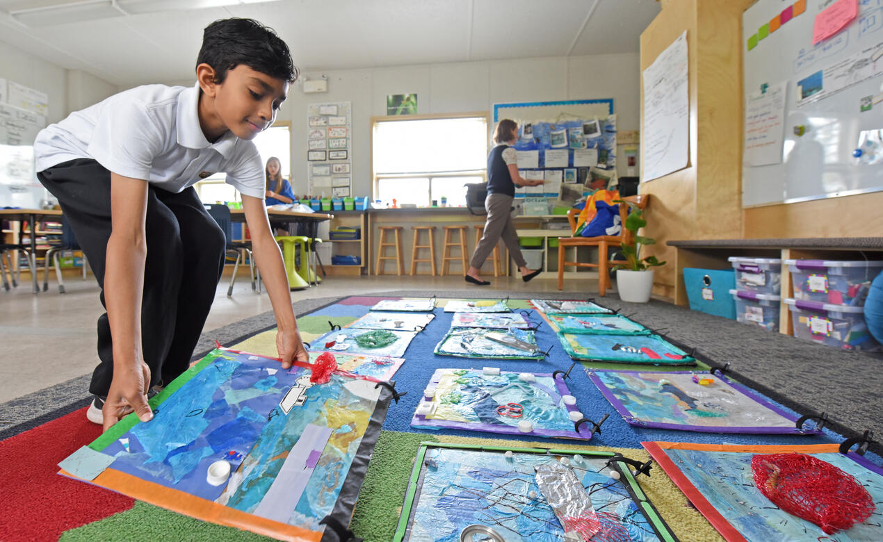 A Grade 5 student places his colourful ocean-themed poster beside all of his classmates' posters on the floor of the classroom