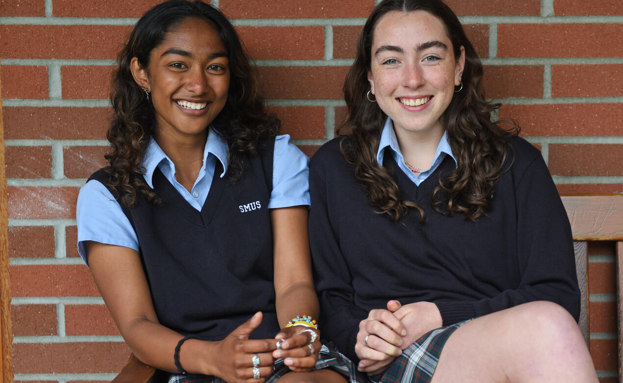 Two SMUS students, Maya and Rowan, sit on a bench in front of a brick wall, smiling for a photo.