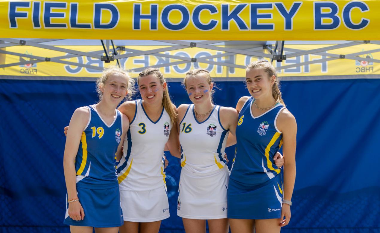 Four SMUS field hockey athletes pose in front of a Field Hockey BC sign.