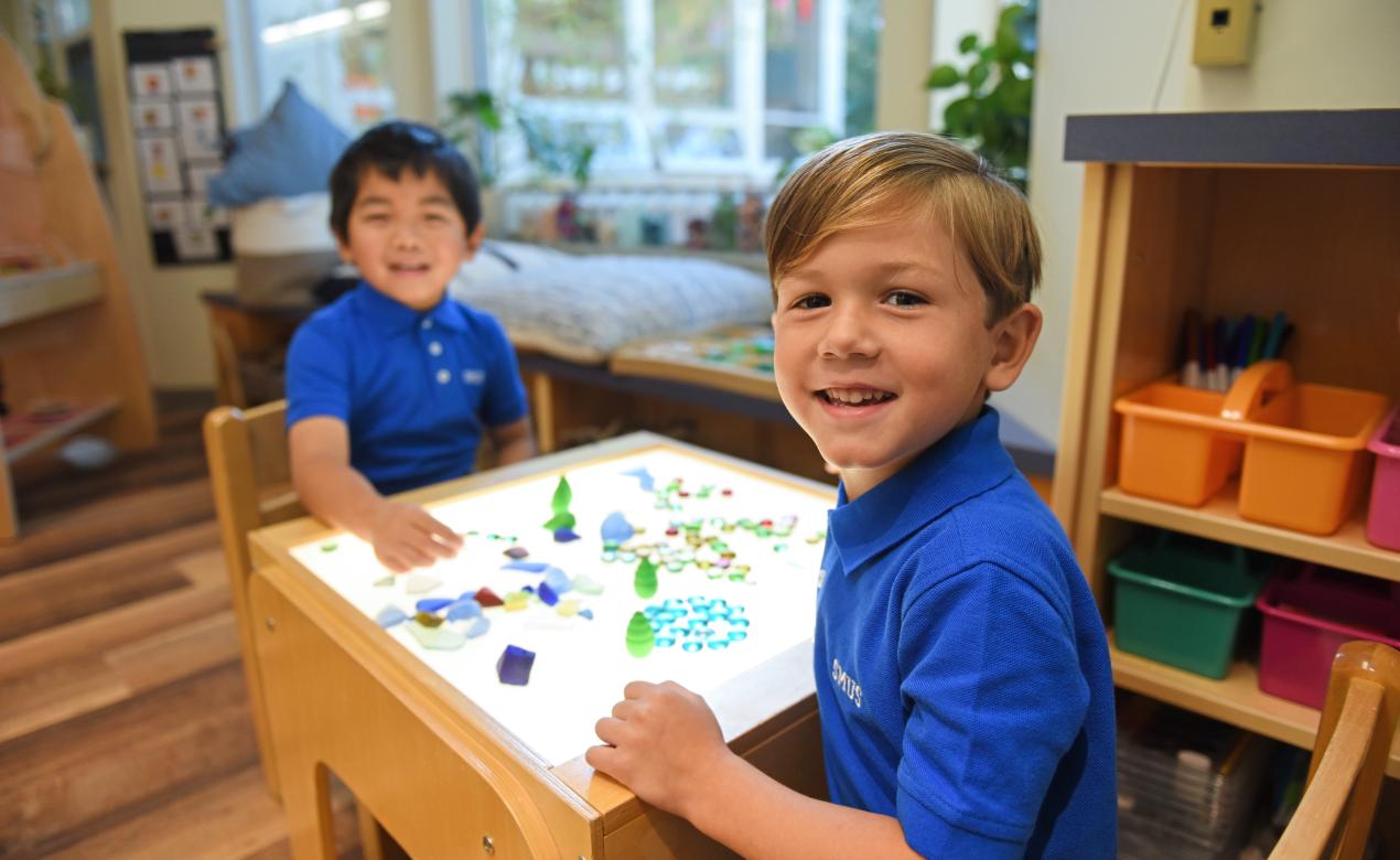 Two Junior Kindergarten students smile at the light table in the classroom