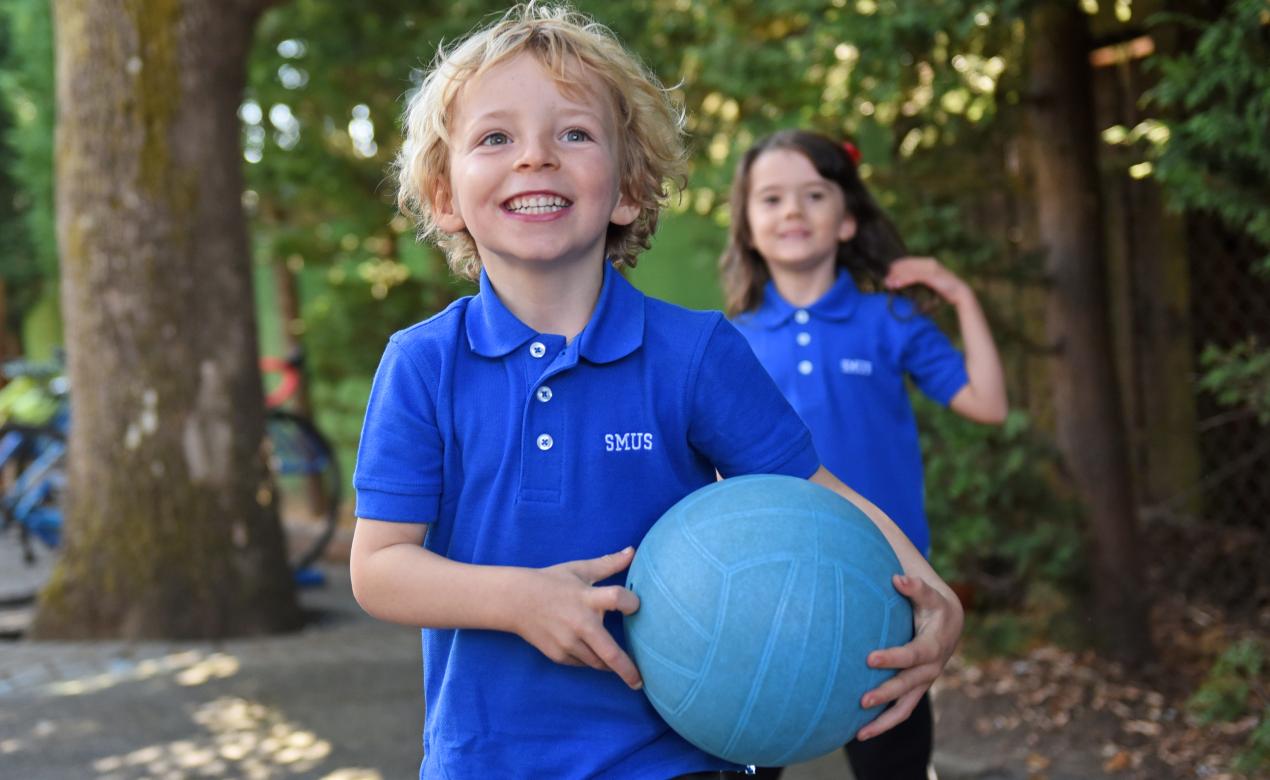 A Junior Kindergarten student smiles while playing outdoors with a ball