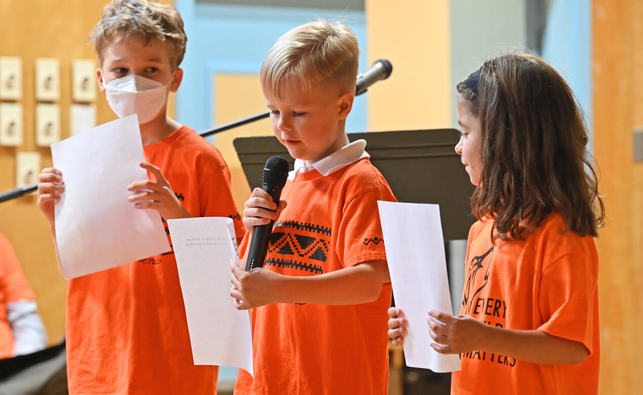Three Junior School students wear orange shirts while speaking during a ceremony in the gym.