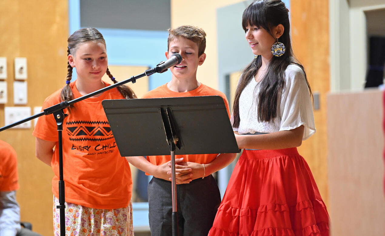 A trio of Middle School students stand at a microphone reading a story during the National Truth and Reconciliation ceremony at the Junior School.