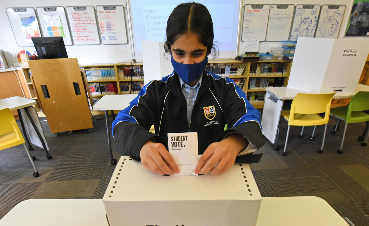 A student wearing a mask inserts a ballot into a ballot box during a mock election in the Middle School library.