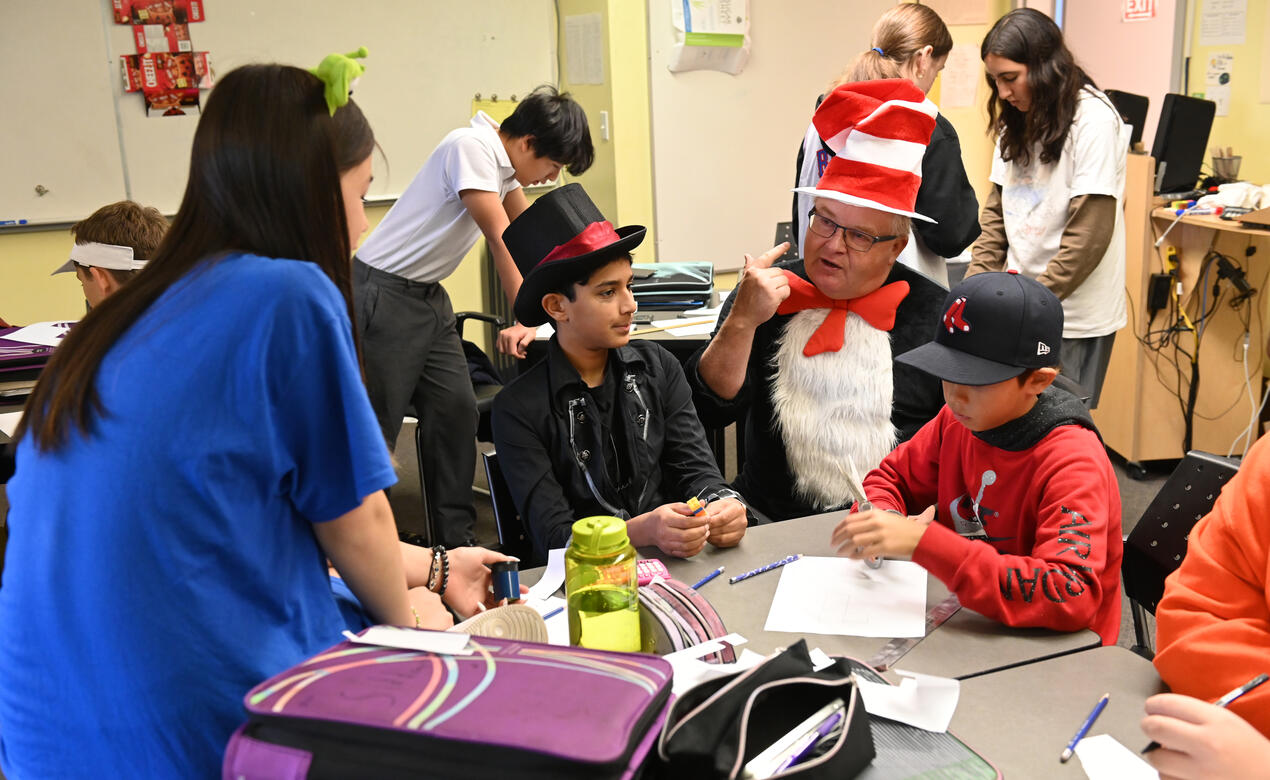 A teacher, dressed as the Cat in the Hat, sits and works with a group of costumed students on Halloween