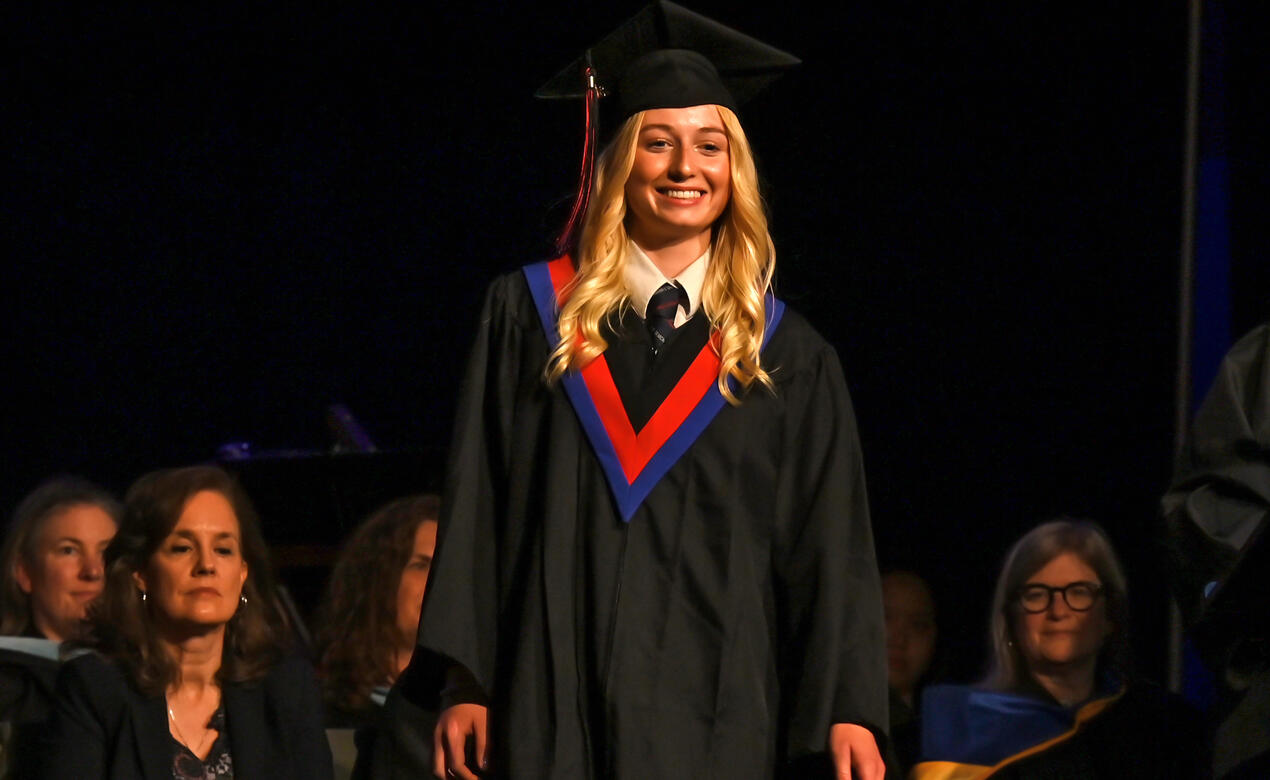 A student wearing cap and gown smiles on stage during graduation