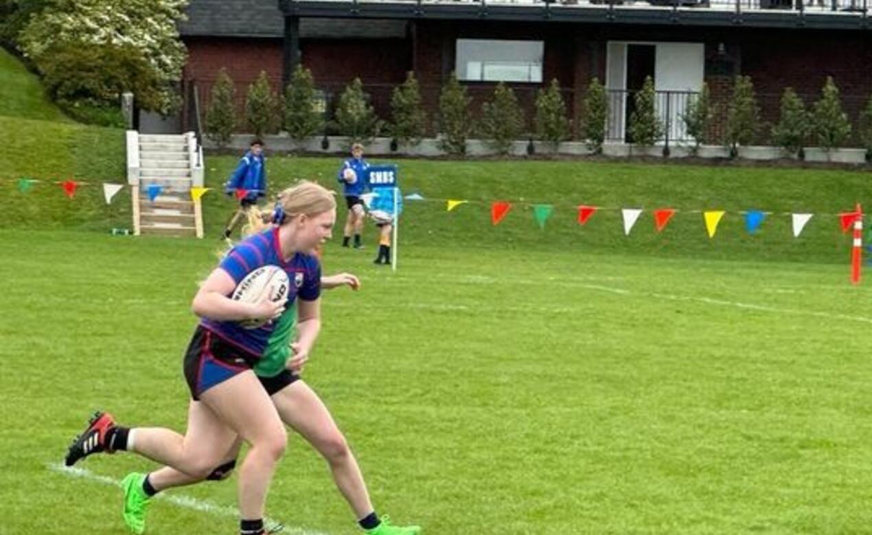 SMUS Rugby player carries ball as an opponent chases her down the grass field as onlookers cheer from Wenman Pavillion.