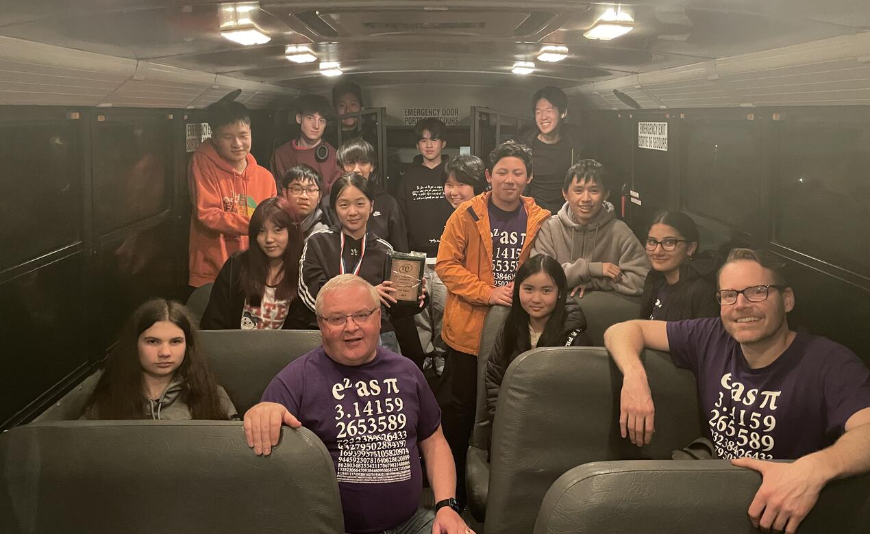 A group of students and their two teachers pose for a group photo from the inside of a bus.