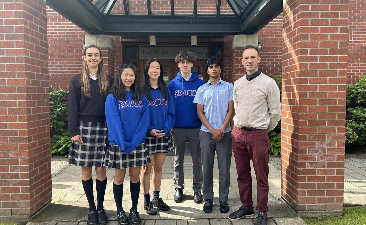 Five students in uniform stand under the brick archway on the SMUS campus along side their teacher