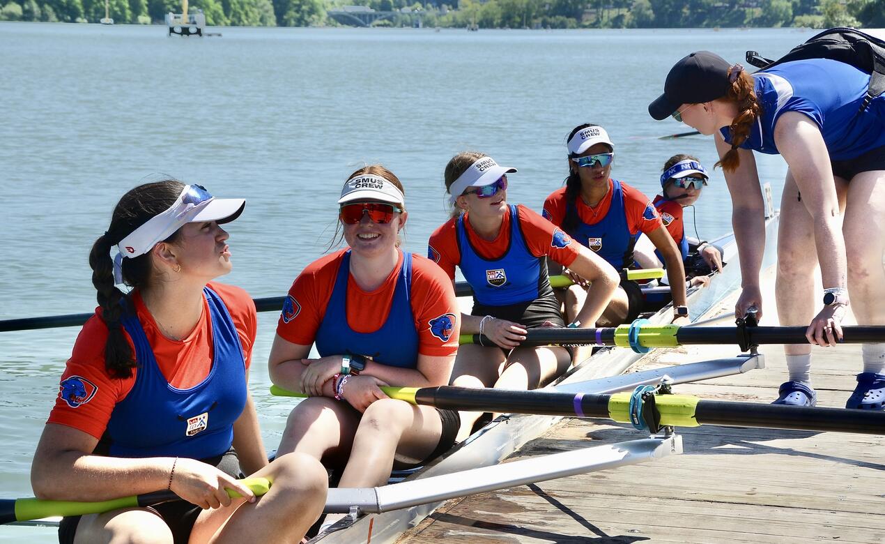 SMUS coxless four women sit in their boat talking to a coach as they prepare for a race.