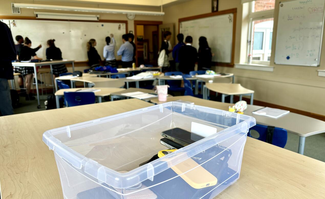 A plastic bin of student-collected cell phones sits on a table while students engage in learning at the whiteboards around the classroom in the background.