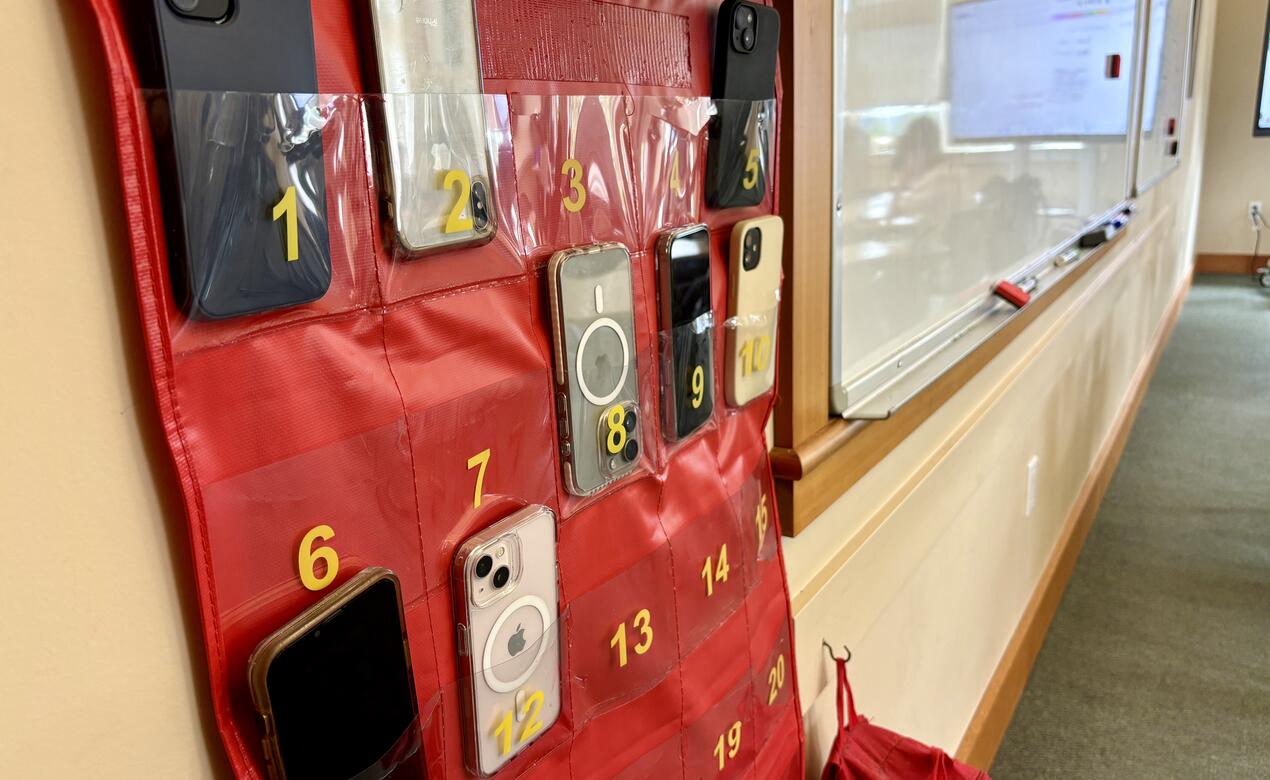 A large folio with numbered pouches holds a variety of student phones hung on the wall of a classroom