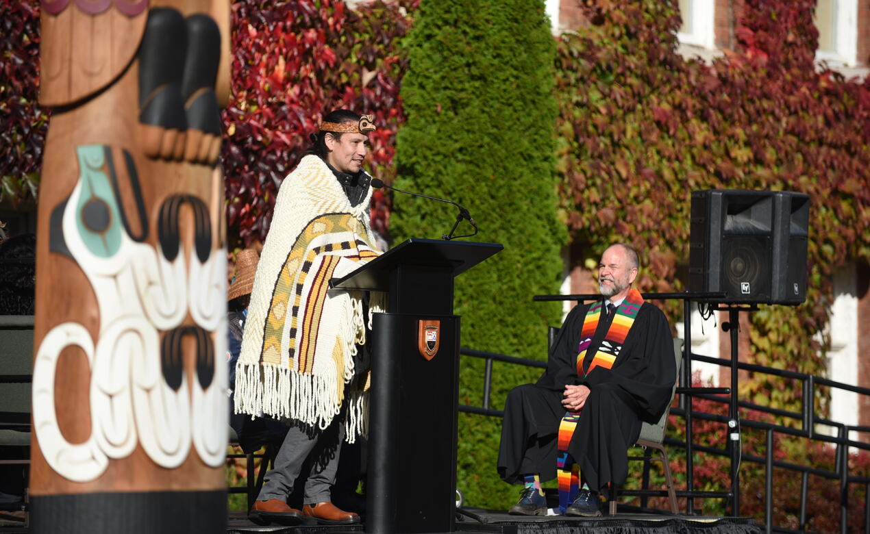 Carver Dylan Thomas addresses the crowd at the podium with his carved Welcome Pole in the foreground