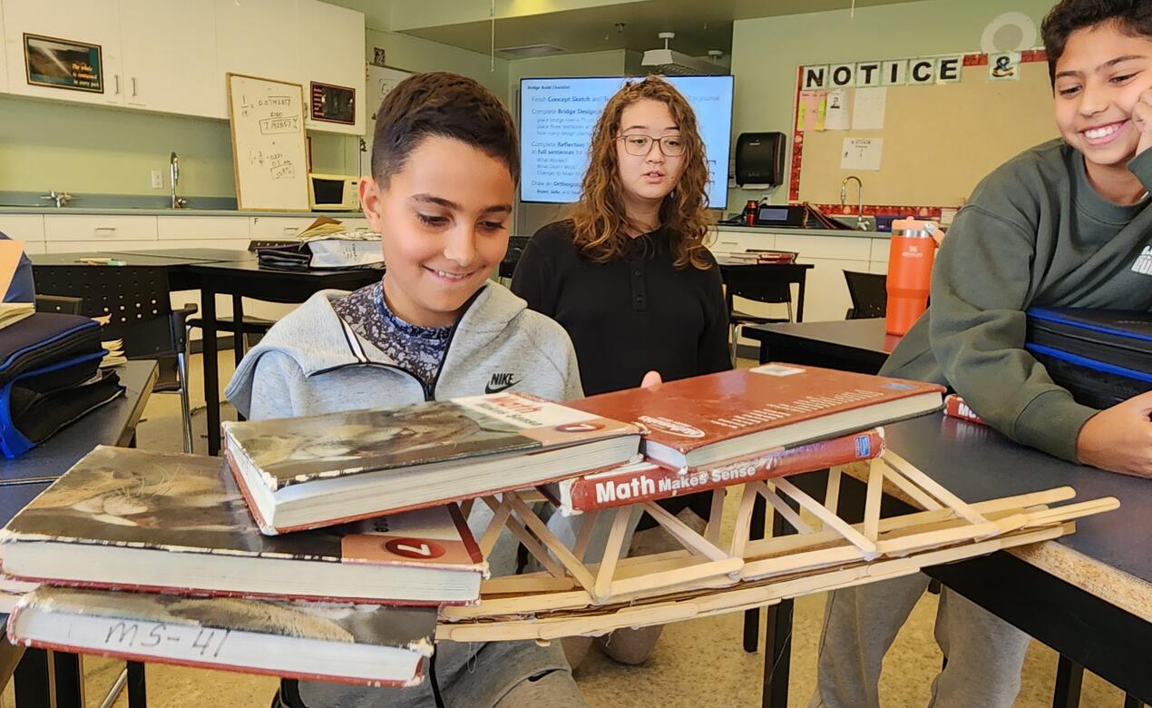 A middle school student watches as more books are piled on top of his bridge to see if it will collapse or not. 