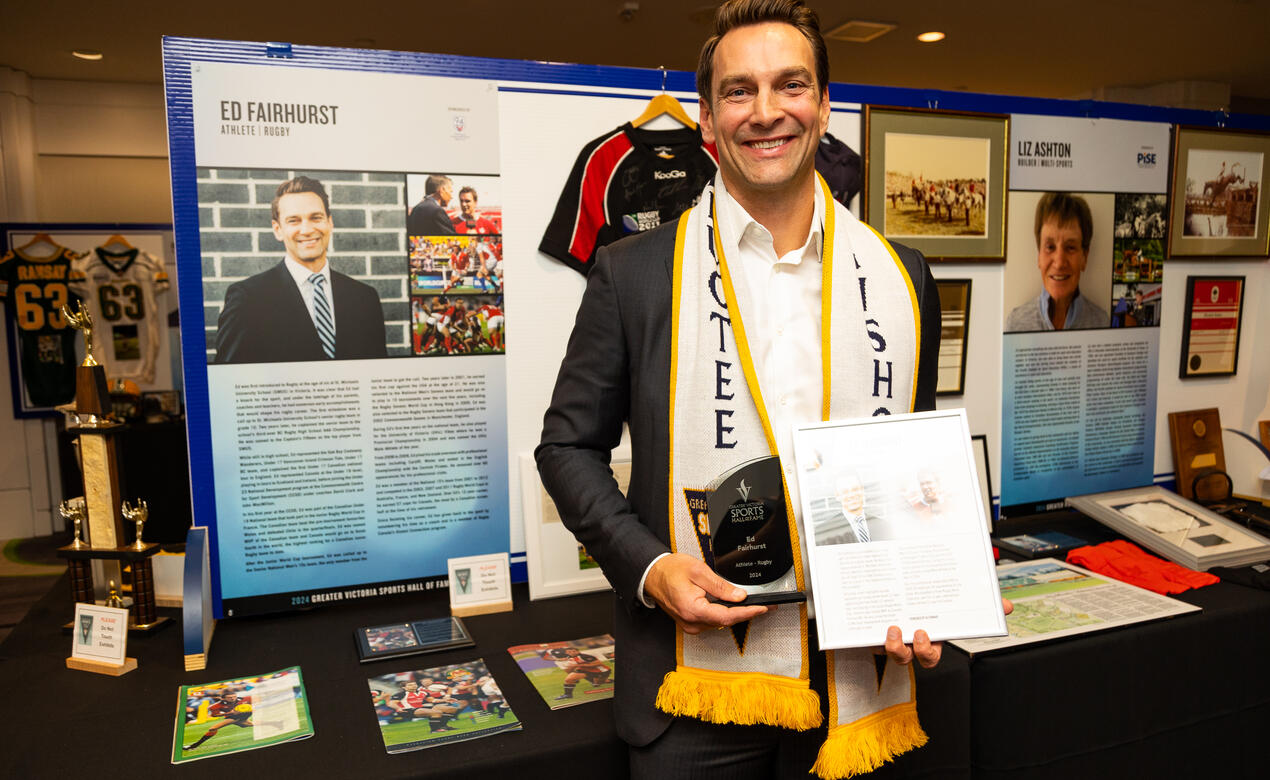 Alumni Ed Fairhurst poses in front of a display of his memorabilia holding his hall of fame induction award