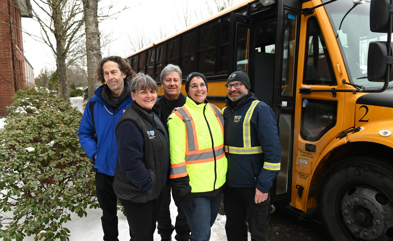 Transportation Manager Jody Bright and four bus drivers smile in front of a bus with snow on the ground. 