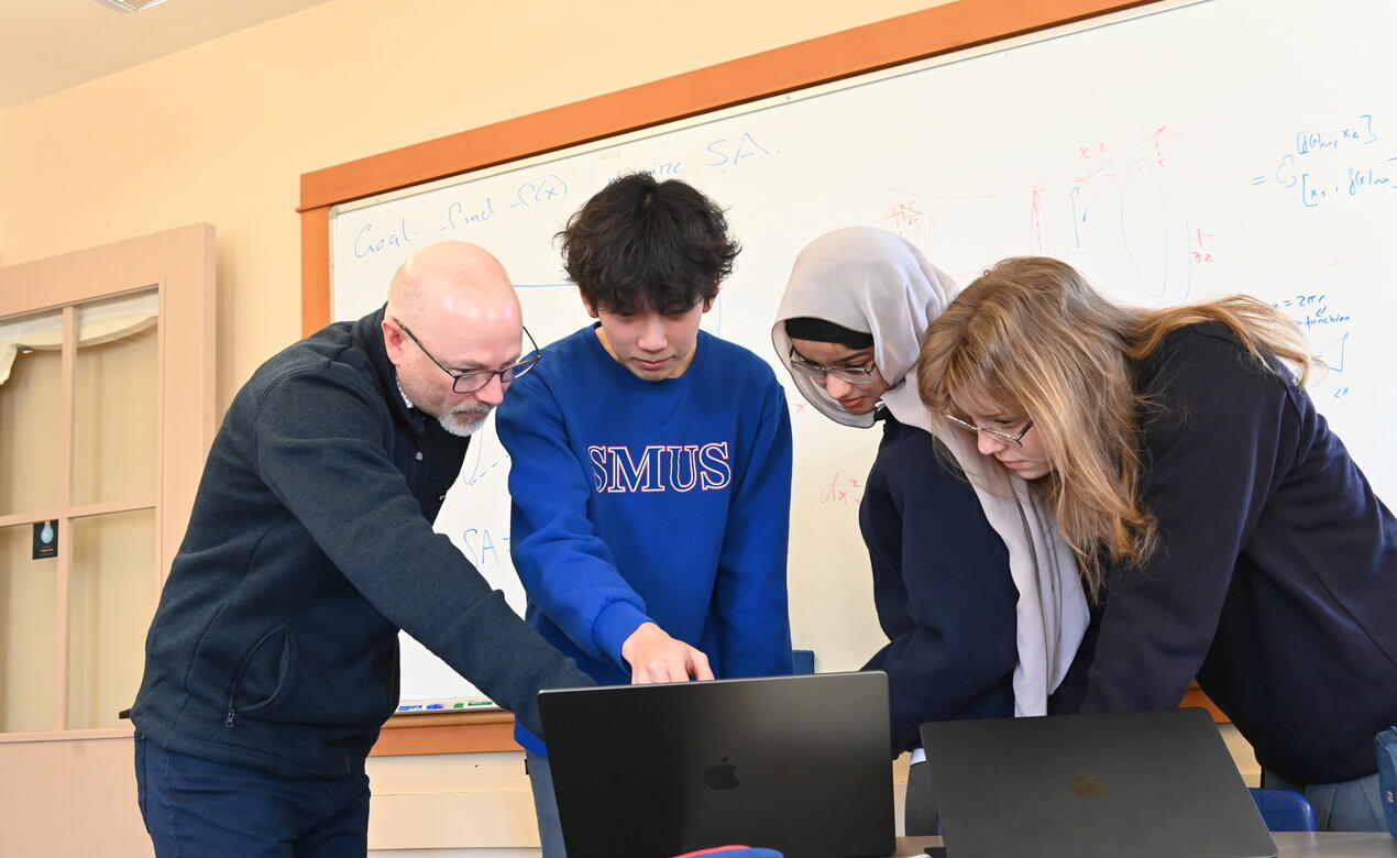 Math teacher Mathew Geddes points to a laptop as he explains something to three SMUS students