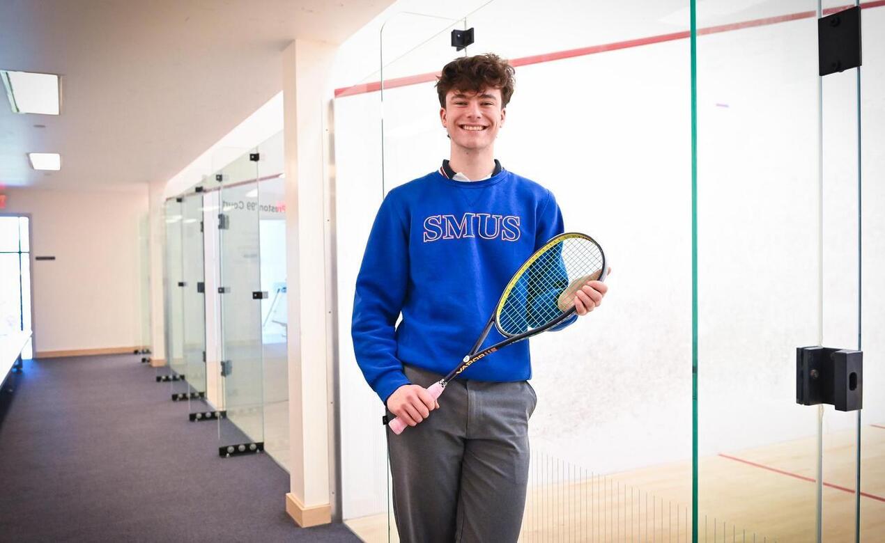 Grade 12 student Anderson Brown leans against the glass of a squash court while holding his racquet and smiling. 