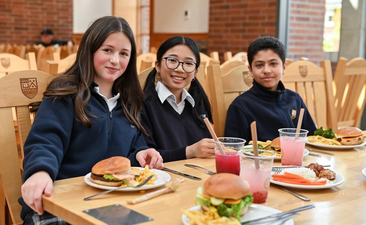 Three Middle School students smile with plates of food in front of them in the Graves Dining Hall