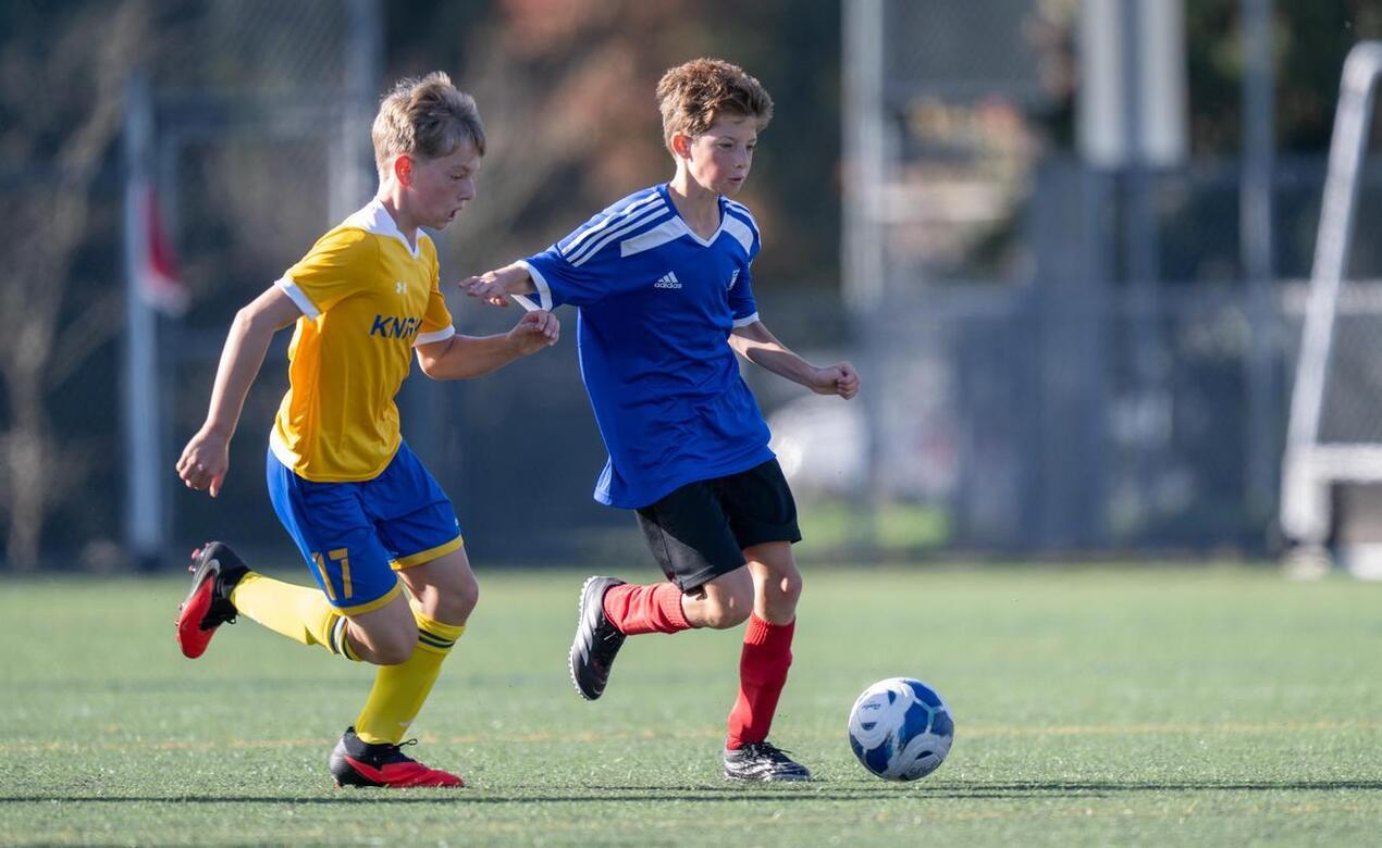 Two boys chasing a soccer ball, one wears a yellow jersey and the other wears a blue jersey. The boy with the blue jersey is about to kick the ball.
