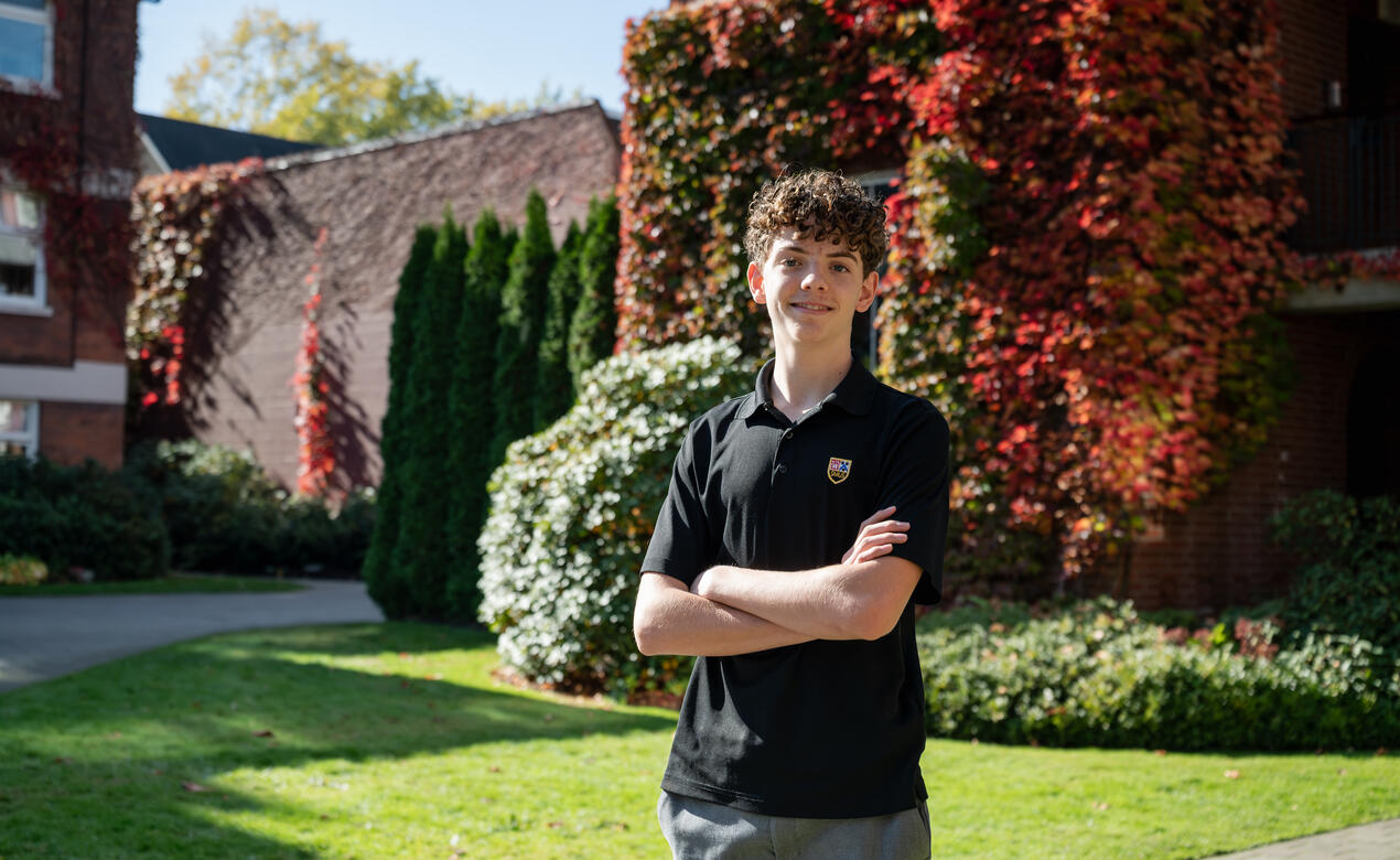 A student in an SMUS polo smiles with arms crossed on a sunny lawn in front of a brick building covered in red autumn ivy.