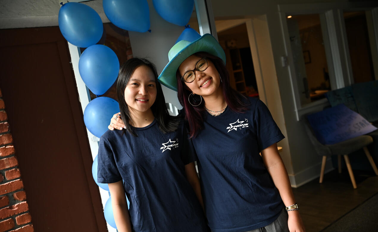Two students in matching navy Symons House T-shirts smile together in a doorway decorated with blue balloons, one wearing a teal cowboy hat.