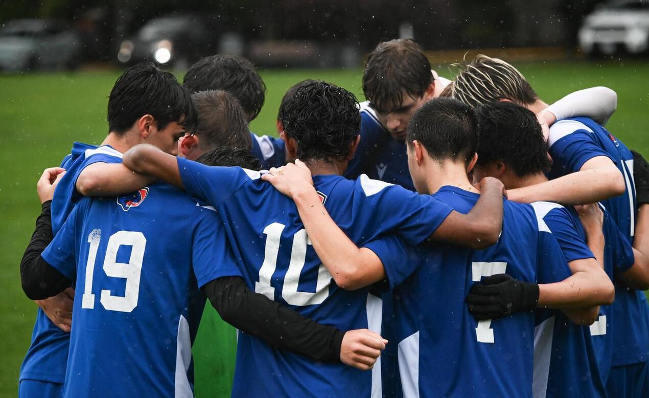 Boys on a soccer team huddled on a field in the rain wearing blue and white jerseys with their heads down and their arms around each others shoulders