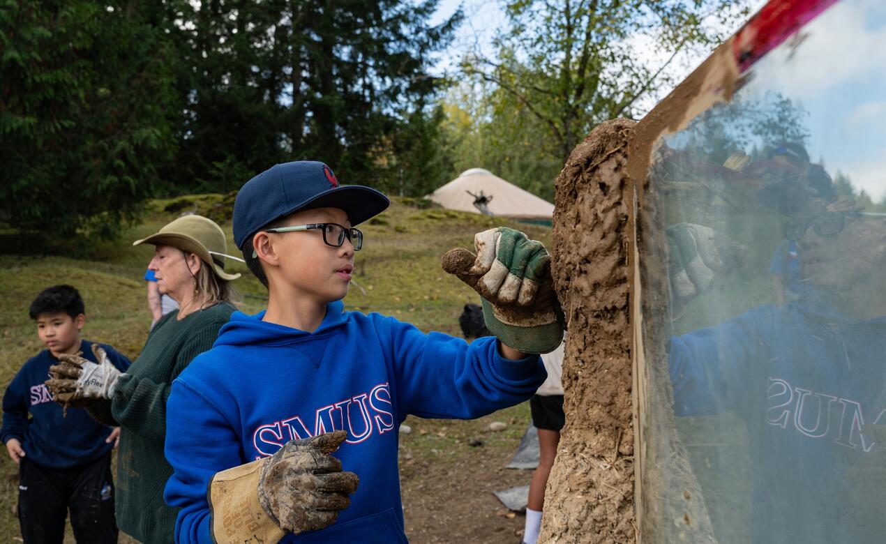 A student with worker gloves on pats down mud on to a wall at EcoVillage