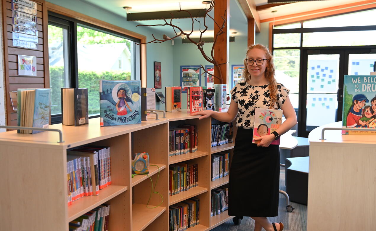Thea Wilson-Scorgie, Junior School Librarian,&nbsp;stands beside a bookshelf displaying picture books in a bright library with large windows.