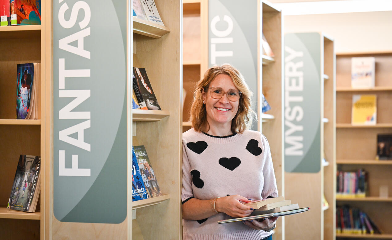 Sarah Bailey, Middle School Librarian, holds a stack of books beside the Fantasy, Realistic and Mystery shelves in the library.