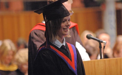 A student wearing cap and gown smiles on stage during graduation.