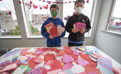 Two students hold up colourful Valentine's Day cards while standing in front of a pile of more cards.