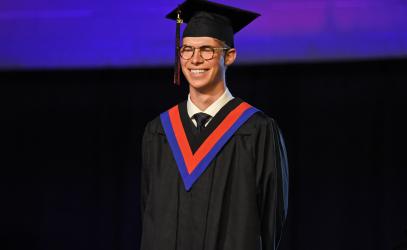 A student wearing cap and gown smiles on stage during graduation.