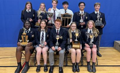 A group of 11 Senior School students pose against a blue backdrop in the gymnasium while holding trophies and plaques.