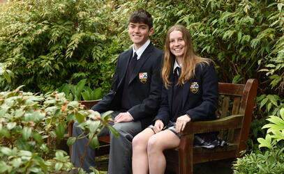 Head Prefects Ryan and Talia pose for a photo while sitting on a wooden bench in front of green flora.