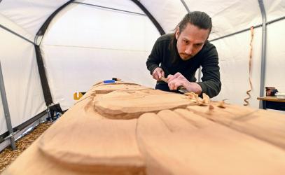 Coast Salish artist Dylan Thomas is crouched over a large wooden post and uses a tool to shave it.