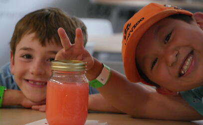 Two smiling children smile while one holds a peace sign with his hands over a jar with a coloured liquid in it on a science day at camp.