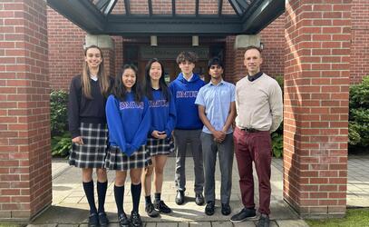 Five students in uniform stand under the brick archway on the SMUS campus along side their teacher