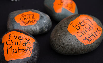 Four rocks lay on a table with orange shirts painted on each one with the word Every Child Matters written in black.