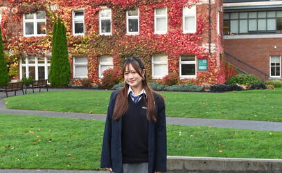 Grade 12 student Yuriko Ishikawa stands and smiles at the camera with the ivy-covered School House Building in the back. 