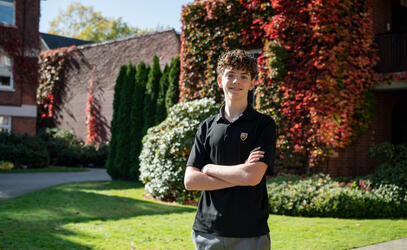 A student in an SMUS polo smiles with arms crossed on a sunny lawn in front of a brick building covered in red autumn ivy.
